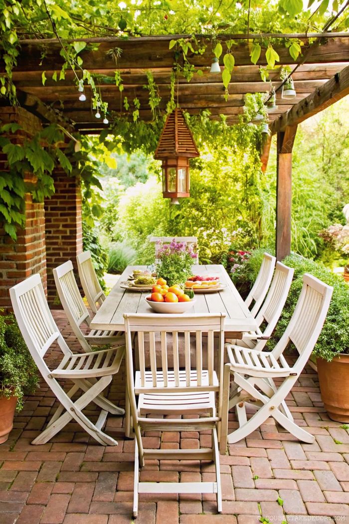 Charming outdoor dining area with wooden table, white chairs, fresh fruits, and greenery cascading from a pergola.