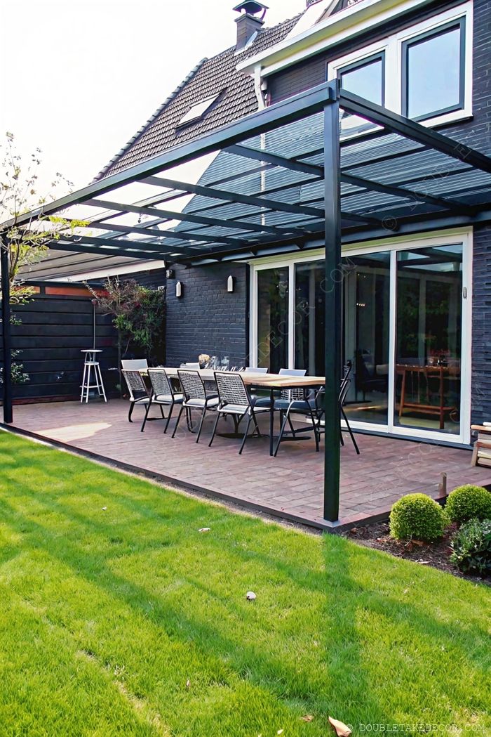 Sleek metal pergola with transparent roof shading a wooden dining table on a paved patio beside a dark brick home with sliding glass doors.