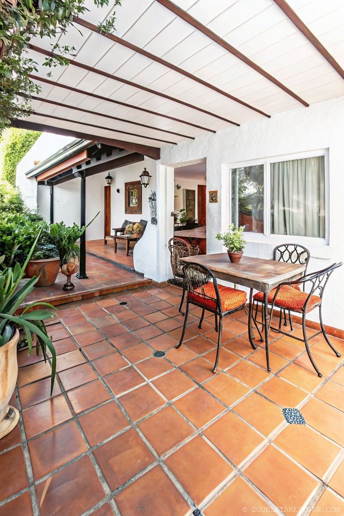 Charming outdoor patio with terracotta tiles, wooden table, wrought iron chairs under a white beamed ceiling cover, surrounded by greenery.