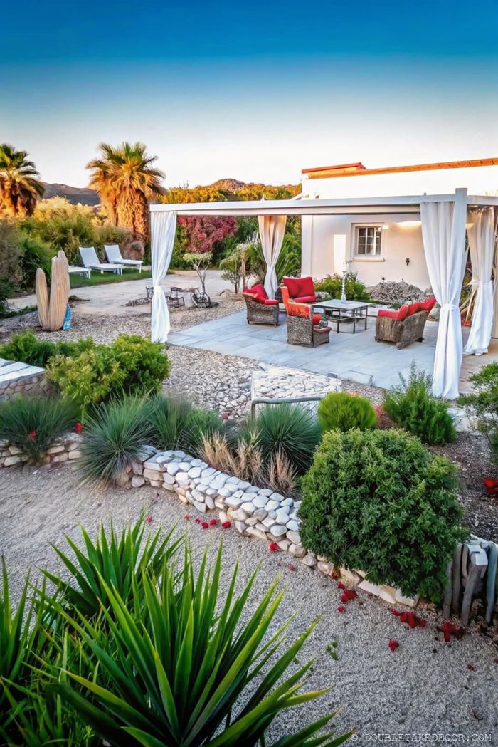 A covered patio with red cushioned seating, white sheer curtains, surrounded by lush greenery and stone pathways in a garden setting.