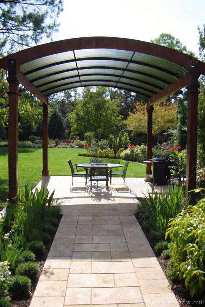 Stylish curved pergola with translucent roof, round dining table and four metal chairs on a stone-paved patio surrounded by lush garden.
