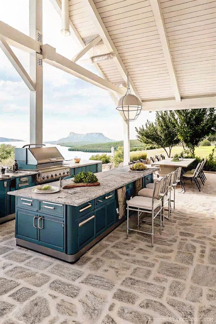 Elegant outdoor kitchen with blue cabinetry, granite countertop, and scenic river views under a white-beamed ceiling.