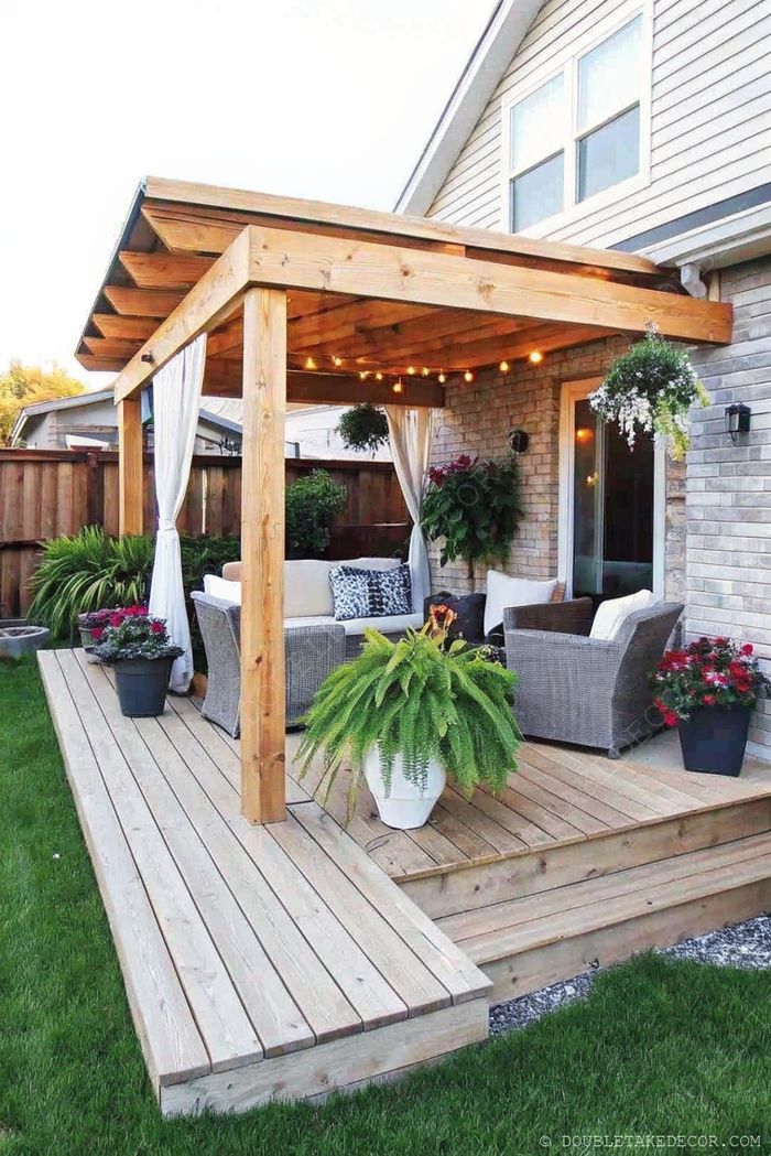 Wooden pergola with white curtains and string lights over comfortable seating on a wooden deck surrounded by greenery.