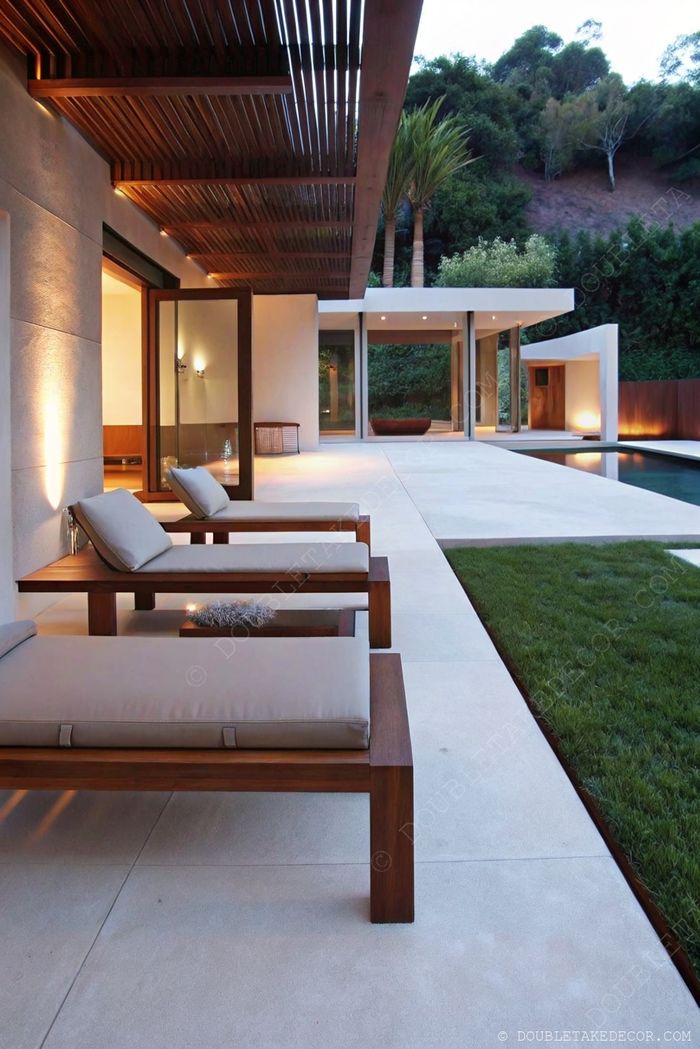 Stylish wooden loungers on concrete patio with pergola, overlooking a landscaped lawn and swimming pool.