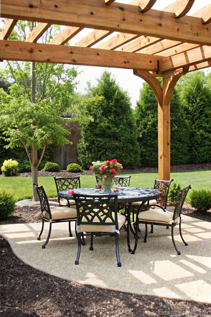 Charming outdoor dining space with wooden pergola, wrought iron table and chairs with beige cushions, surrounded by lush greenery.