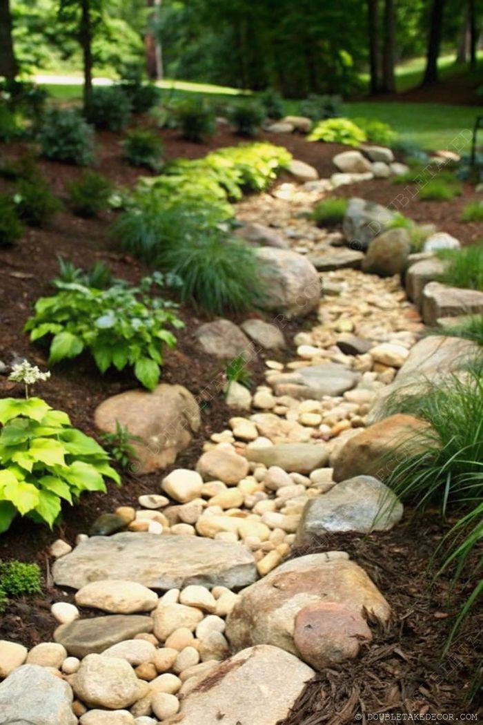 Landscaped garden path with smooth stones forming a dry streambed, bordered by rocks and lush greenery in dappled sunlight.