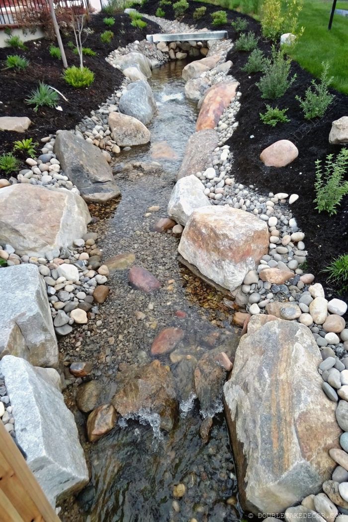 Serene landscape with winding stone-lined stream, flowing over rocky bed, surrounded by lush greenery and dark mulch.