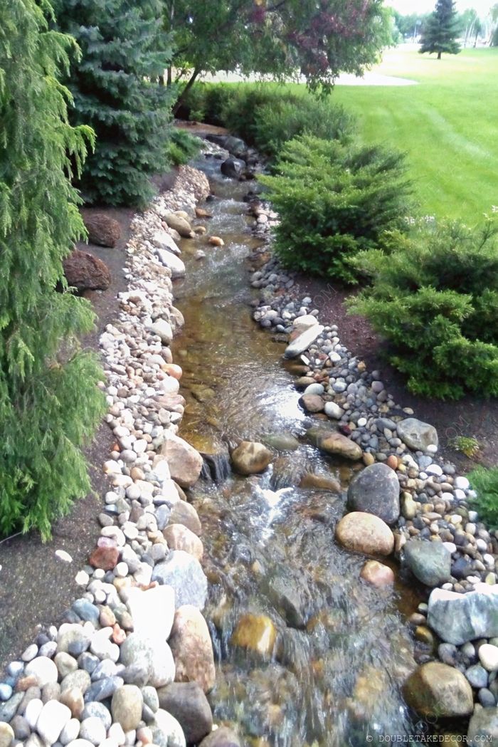 A peaceful landscape with a flowing stream bordered by smooth stones, surrounded by lush greenery and reflecting foliage.