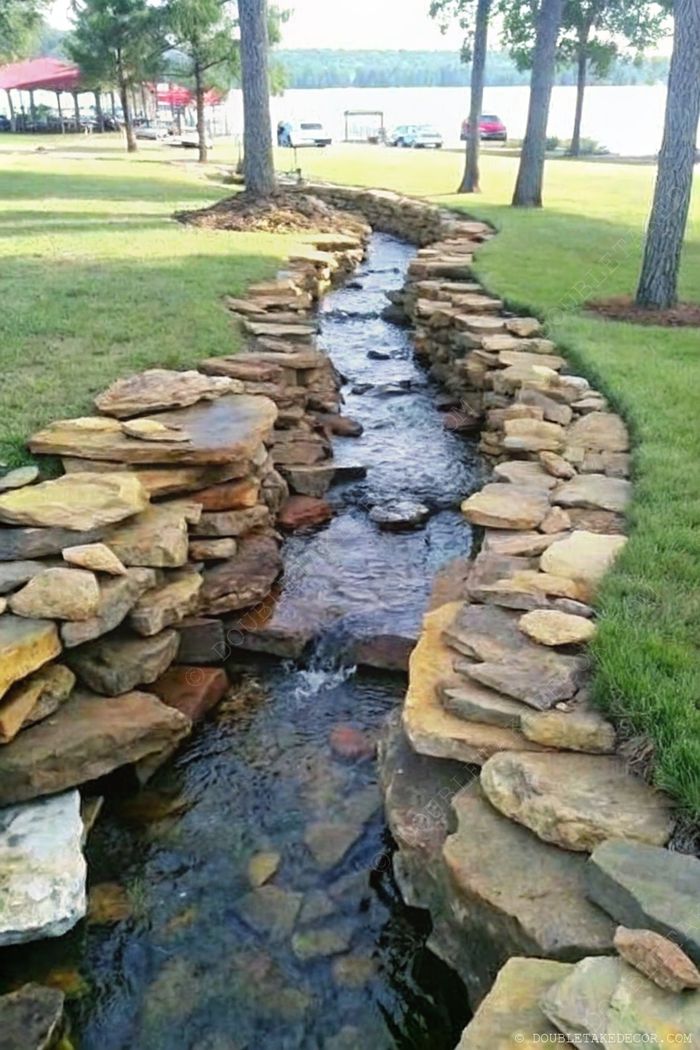 A gentle stream flows through a lawn, bordered by arranged rocks, with lush greenery and trees creating a tranquil rainscaping feature.