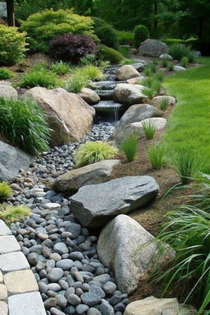 Tranquil garden landscape featuring a gentle stream flowing over smooth stones, surrounded by textured rocks and lush greenery.