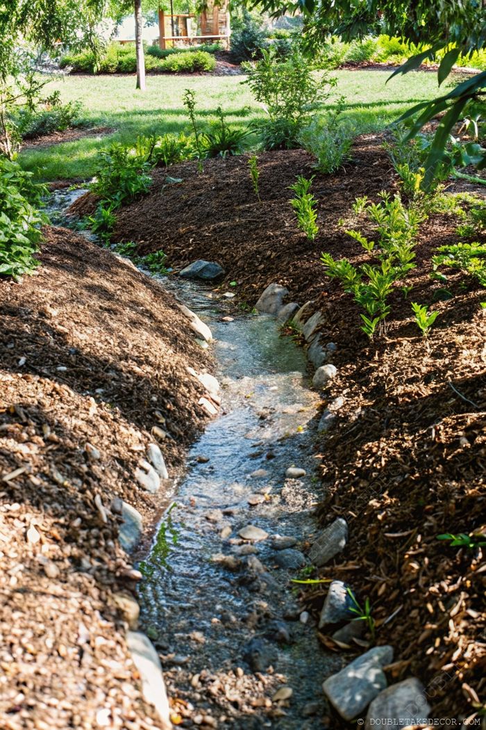 A peaceful backyard stream with smooth stones flowing between mulched banks and lush green vegetation in a harmonious natural setting.