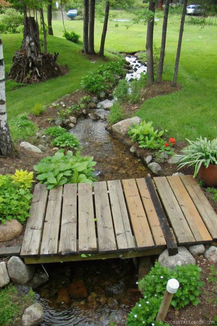 Tranquil garden stream with rustic wooden bridge, surrounded by lush greenery, colorful flowers, and decorative stones.