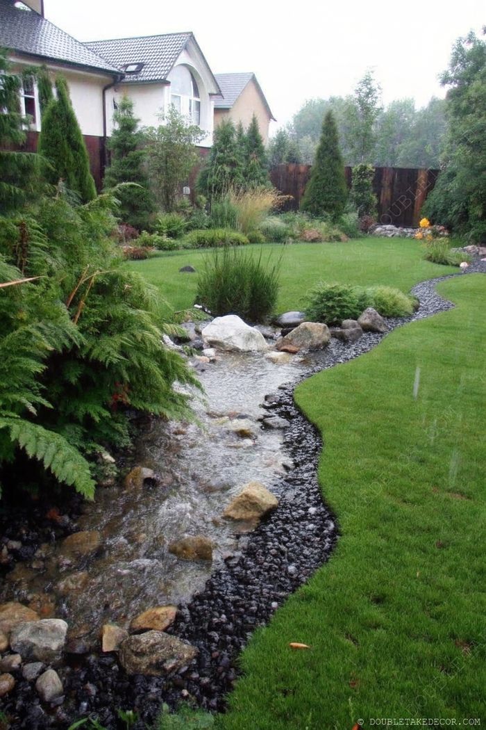 A winding stream with dark stone borders flows through a lush garden with vibrant plants and shrubs under a cloudy sky.