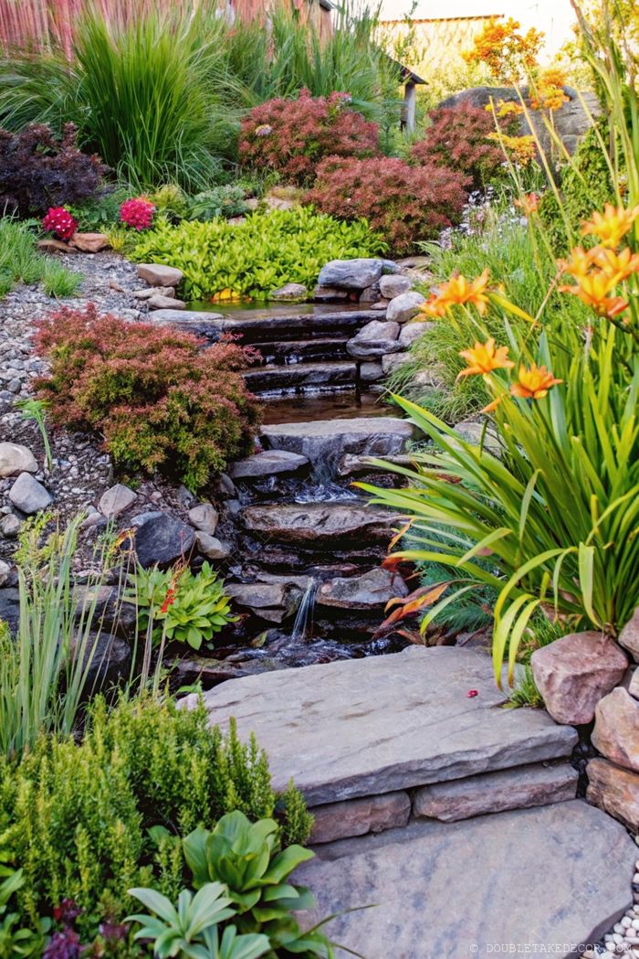 A serene garden waterscape with stone steps flowing alongside a gentle waterfall, surrounded by vibrant flowers and lush greenery.