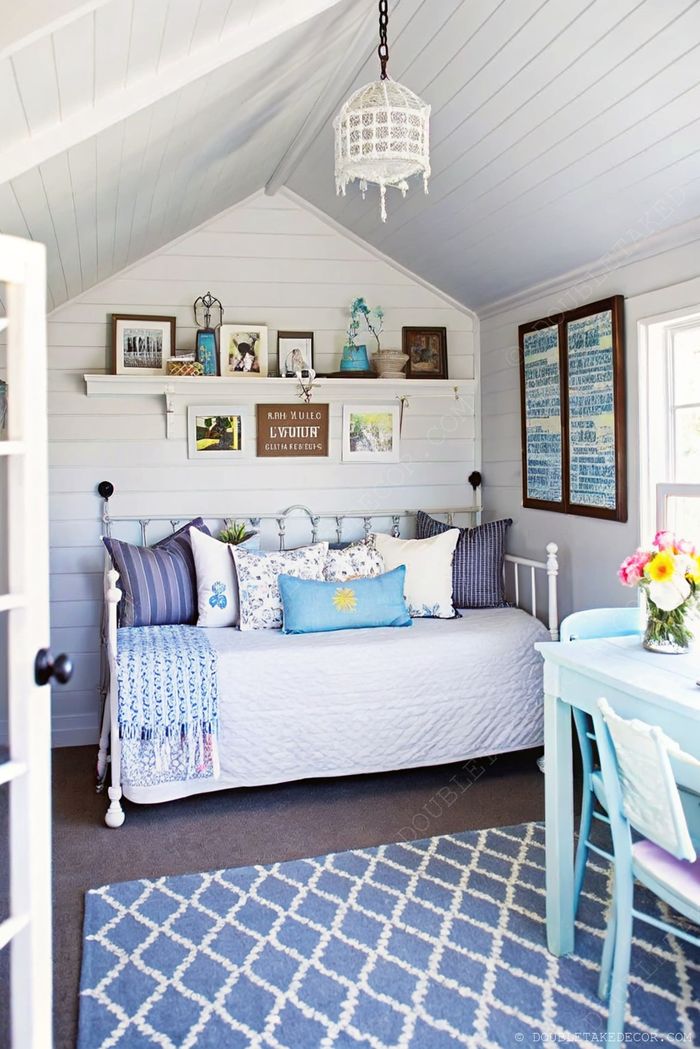 A charming she shed interior with a vintage daybed adorned with colorful pillows, light decor, and a small blue dining table with fresh flowers.