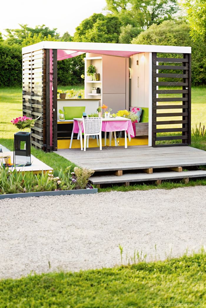 A contemporary she shed with wooden slats and sleek roof featuring a green bench, pink-clothed table, and indoor plants in a serene garden setting.
