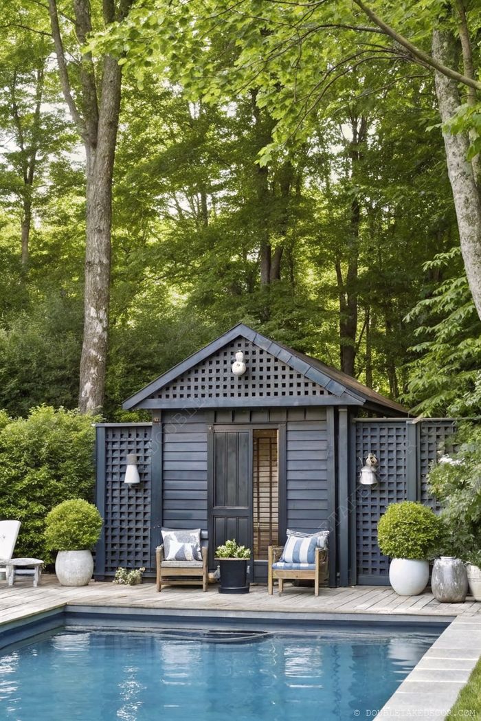 Dark blue she shed with gabled roof and lattice detail, featuring a wooden deck with chairs overlooking a serene swimming pool