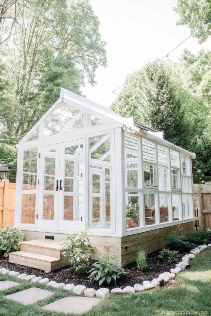 Charming white-framed greenhouse with glass panels nestled in a lush garden with stone pathway and wooden steps