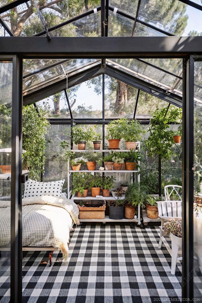 A glass-roofed greenhouse converted into a cozy bedroom with striped bedding, plant-filled shelves, and checkered flooring.