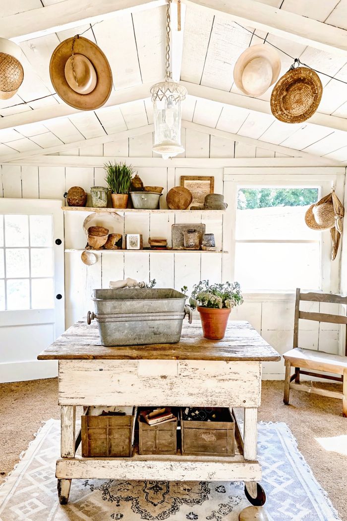 Cozy rustic interior with wooden table, vintage basin, white walls, and overhead shelving displaying gardening items and decorative elements.