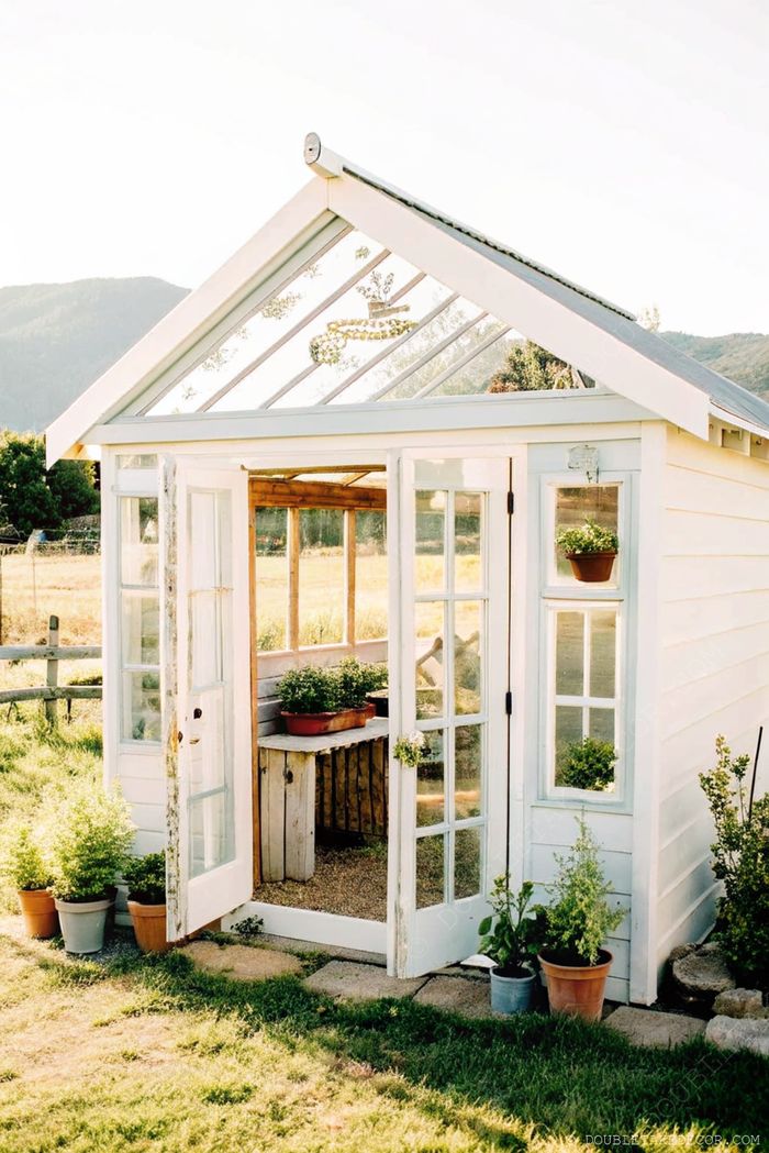 A charming white wooden greenhouse with glass panes, double doors, and a sloped transparent roof, surrounded by potted plants in a serene countryside setting.