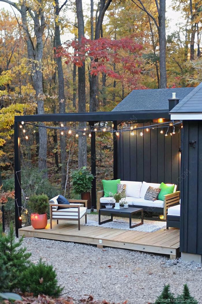 Cozy outdoor deck with white sectional under black pergola adorned with fairy lights, surrounded by vibrant autumn foliage.
