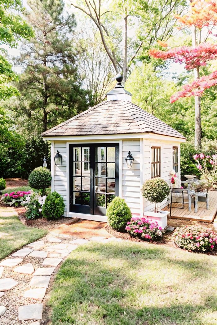 Charming white garden shed with black accents, double doors with windows, surrounded by manicured flower beds and a stone path leading to entrance.