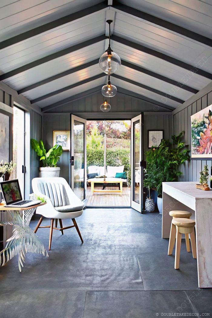 Stylish indoor she shed with gray walls, pendant lights, workspace with laptop, and double doors opening to a lush outdoor area.