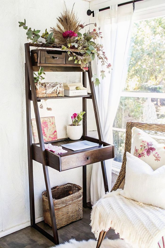 Wooden leaning shelf with plants, books, and decor items, featuring storage drawer and woven basket, beside a cozy chair with throw and floral pillows.
