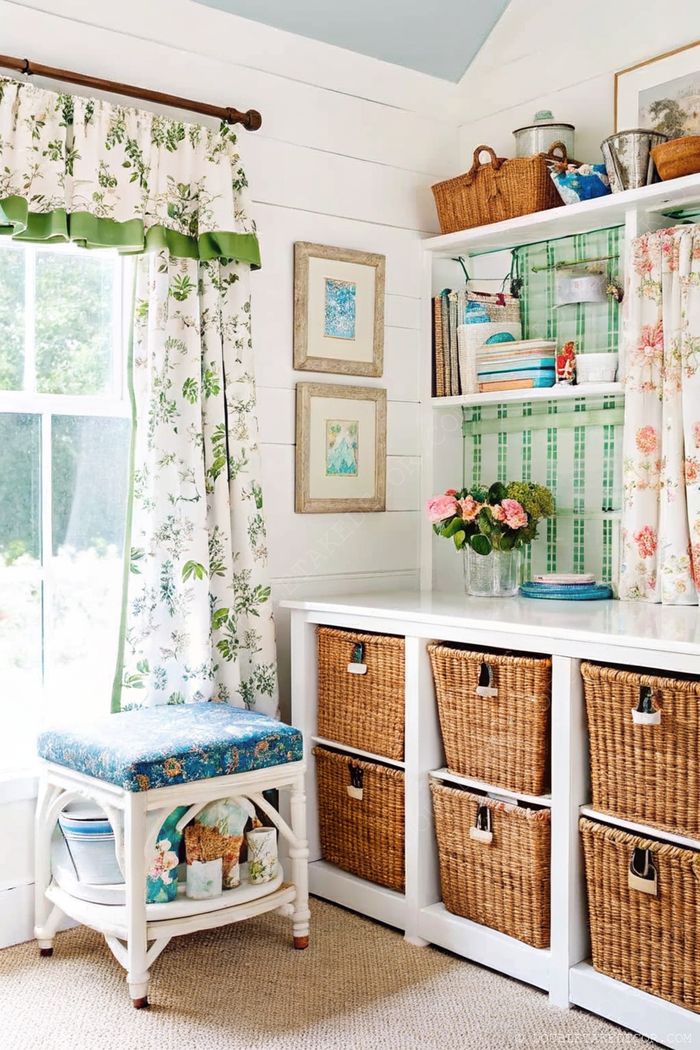A bright she shed interior with floral curtains, blue stool, and white shelving unit featuring organized craft supplies and labeled storage baskets.