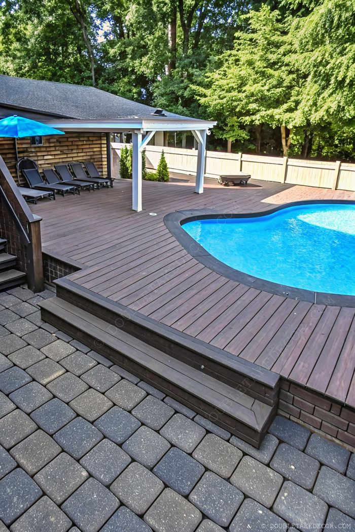 Inviting backyard pool with curved design, wooden deck, black loungers, blue umbrella, and lush green surroundings