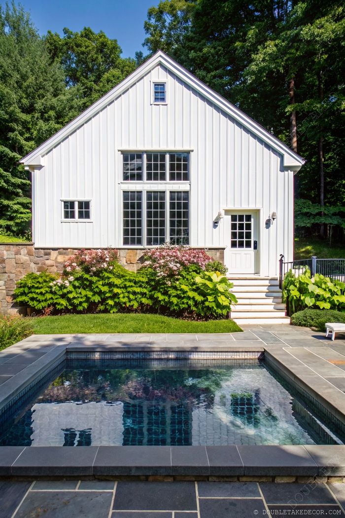 Modern white New England barn-style home with a sleek pool reflecting the architecture, surrounded by slate tiles and lush landscaping.