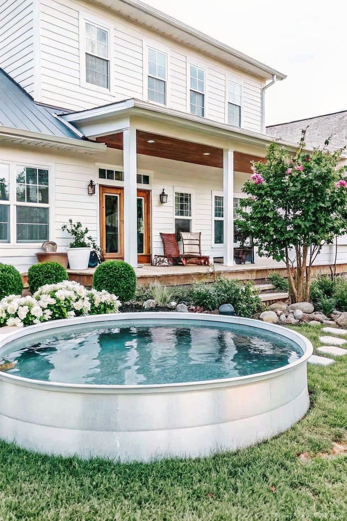 A contemporary circular metal pool adorns a green backyard with stone pathways, white-sided house, and comfortable porch seating.