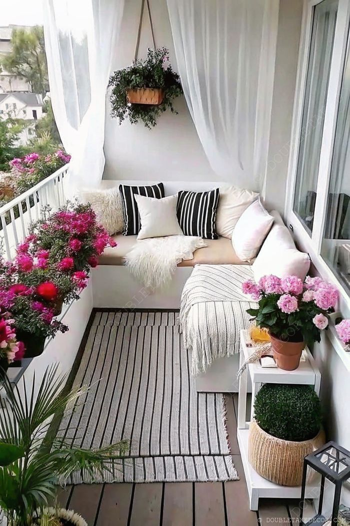 Inviting balcony with comfortable seating, decorative pillows, pink flowers, and sheer curtains creating a serene outdoor sanctuary.