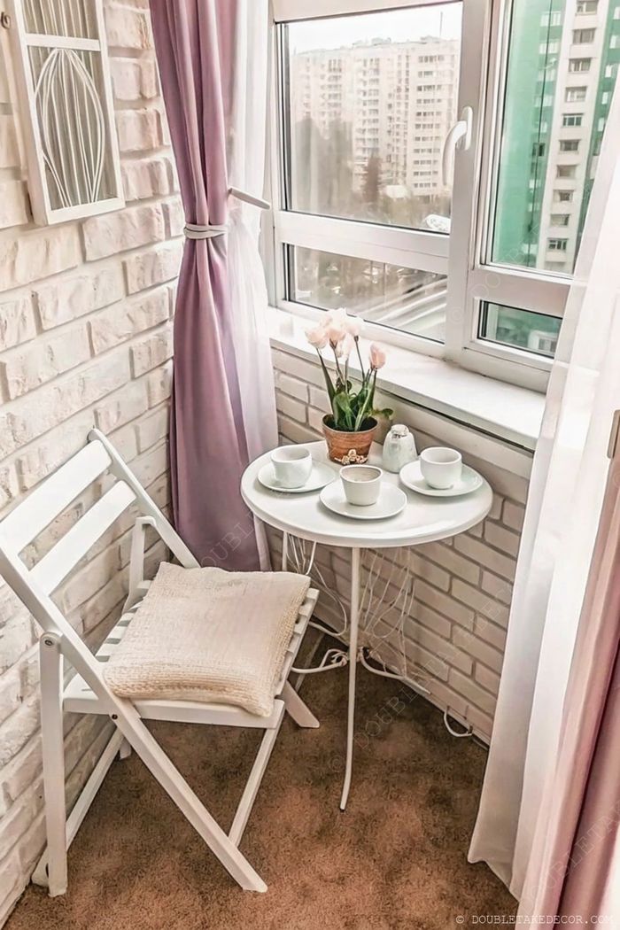 Cozy balcony corner with brick walls, round white table set for tea, pink potted flowers, and a comfortable chair with soft cushioning.