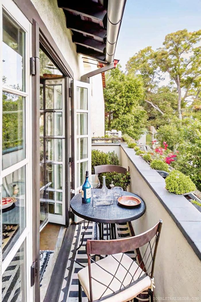 Cozy balcony with round black table, stylish chairs, wine setting, surrounded by greenery and accessible through French doors.