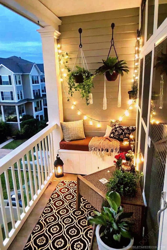 A serene balcony featuring a cushioned bench with pillows, hanging plants, string lights, a patterned rug, and decorative lantern.