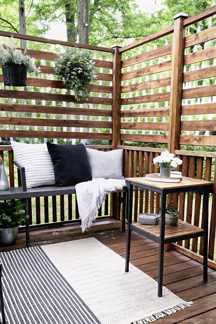 Wooden bench with cushions and throw blanket beside a small table with flowers, surrounded by plants and privacy slats
