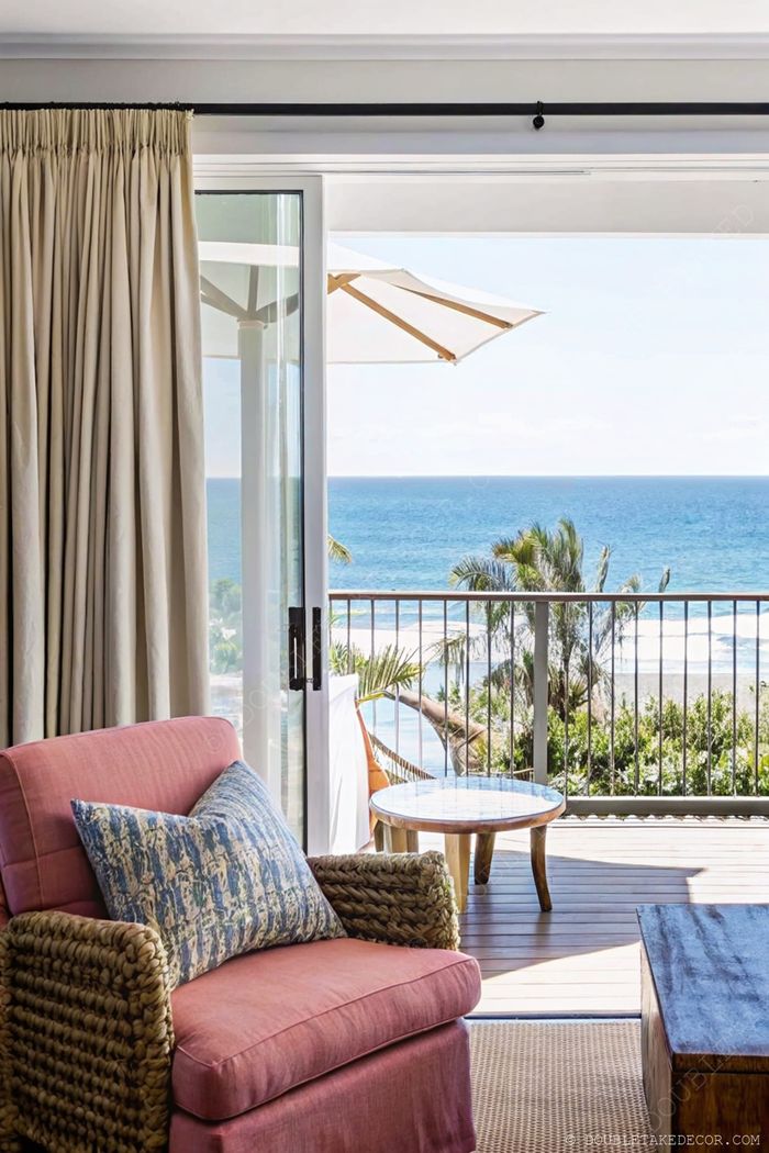 Coral armchair with wicker arms and decorative pillow near glass balcony door with ocean view and outdoor seating area.