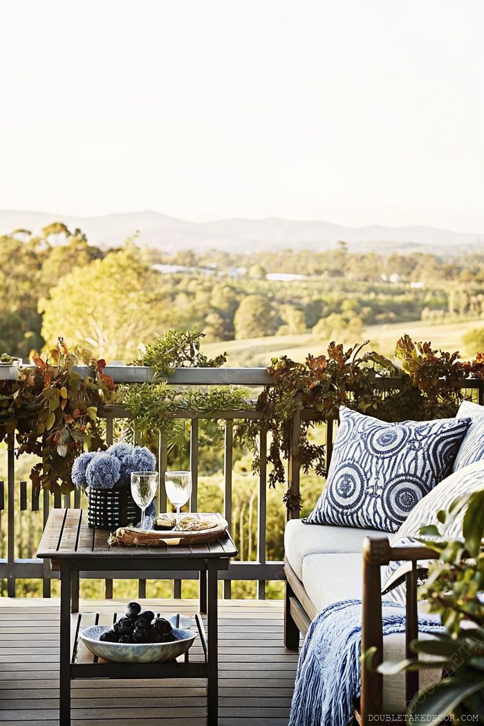 Cozy balcony retreat with wooden table, wine setup, and comfortable seating overlooking scenic mountain views through lush railing greenery.