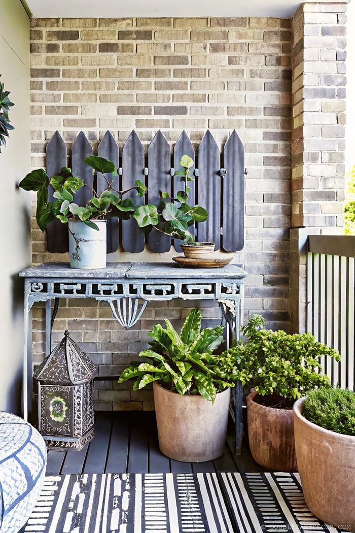 A tranquil balcony garden featuring a blue ornate table with potted plants against a brick wall, complemented by a black fence panel and striped rug.