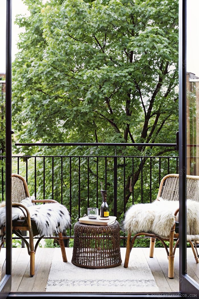 Intimate balcony with two woven chairs, fur throws, and a side table on wooden decking, framed by lush greenery through glass doors.