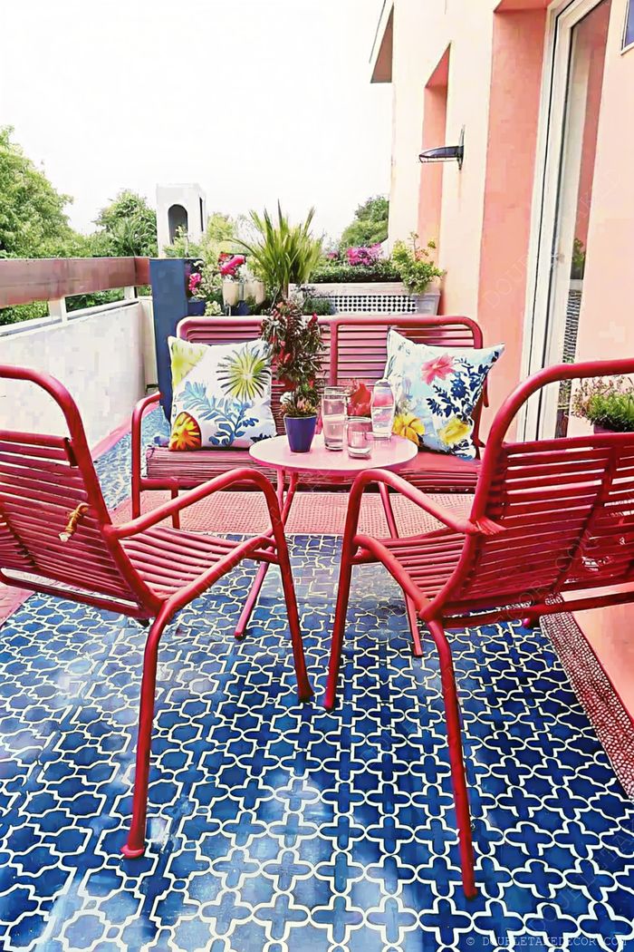 Cozy balcony with red metal chairs, round table, geometric blue tiled floor, and lush potted plants creating a colorful outdoor retreat.