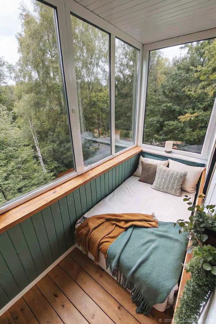A serene enclosed balcony with large windows, featuring a daybed with neutral pillows, colorful throws, green walls, and potted plants creating a peaceful retreat.