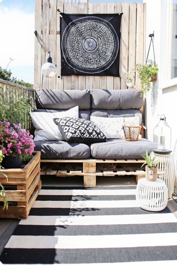 Cozy balcony retreat featuring a gray pallet couch with patterned cushions, hanging plants, and a striped rug creating an inviting outdoor space