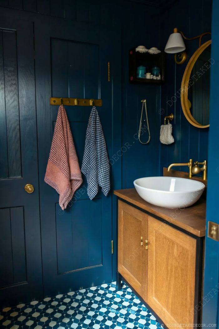 Salle de bain élégante bleu marine avec touches de bois, serviettes corail et bleu à carreaux sur crochets en laiton, miroir rond au-dessus d’une vasque blanche.