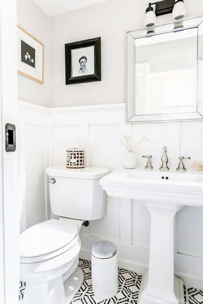 Salle de bain lumineuse avec panneaux blancs, lavabo sur pied, œuvres encadrées et sol graphique noir et blanc.