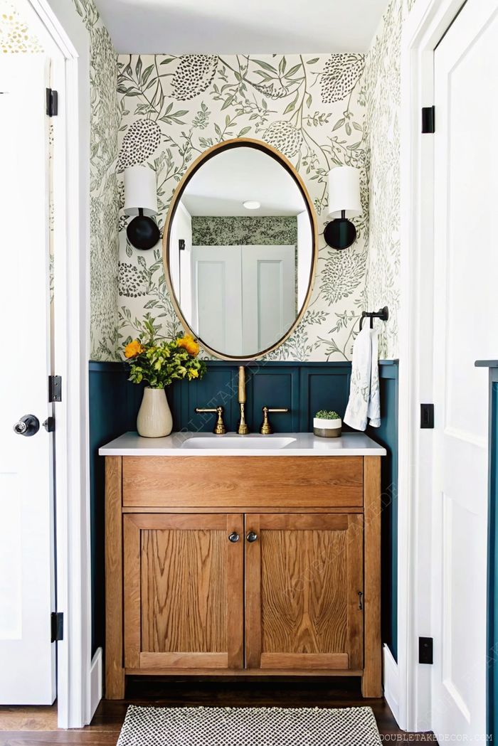 Petit cabinet de toilette avec papier peint fleuri, miroir rond doré, plan blanc et meuble en chêne avec touches de noir.