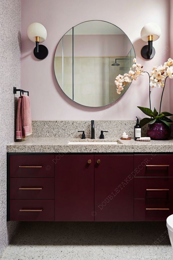 Salle de bain contemporaine à murs roses, meuble bordeaux à poignées bronze, miroir rond et jolies appliques murales.