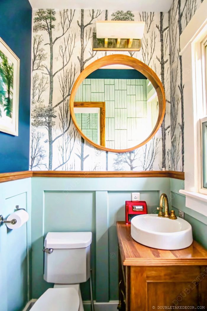Petit espace déco avec papier peint à motif arbres, mur bleu marine, miroir rond en bois, lavabo blanc et meuble sous vasque en bois chaud.